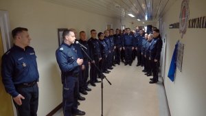 Fotografia: od lewej strony na fotografii grupa kobiet i mężczyzn w granatowych mundurach policyjnych  przed ściną z logo Laboratoriom Kryminalistycznego Komendy Stołecznej Policji pod którym znajduje się tablica upamiętniająca sześć psów policyjnych, które zginęły tragicznie 21 lutego 2020 roku.