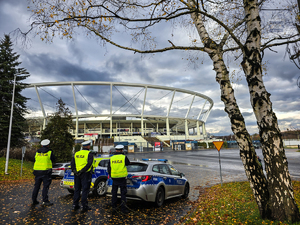 Trzech umundurowanych policjantów ruchu drogowego oraz radiowóz na tle stadiony zabezpieczających ćwiczenia