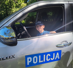 Chłopiec w czapce policyjnej siedzi za kierownicą pojazdu służbowego Centralnego Laboratorium Kryminalistycznego Policji.