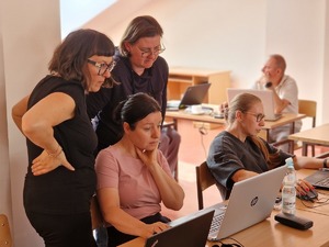 Zdjęcie przedstawia uczestników warsztatów. dwie osoby siedzą przed komputerami inne dwie stoją i realizują zadania zlecone przez prowadzącego.