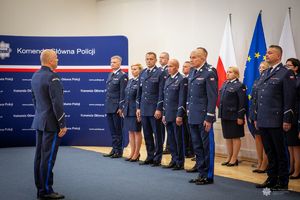 Zdjęcie przedstawia uroczystość w Komendzie Głównej Policji w Polsce. Widzimy grupę umundurowanych funkcjonariuszy Policji ustawionych w szeregu, prawdopodobnie podczas ceremonii wręczenia awansów, odznaczeń lub ślubowania. Przed nimi stoi inny funkcjonariusz, również w pełnym mundurze galowym, który prawdopodobnie pełni funkcję prowadzącego uroczystość lub komendanta.Tło stanowi niebieska plansza z napisem „Komenda Główna Policji” oraz logotypami i adresami stron internetowych Policji (m.in. www.cikp.policja.pl). Po prawej stronie widoczne są flagi: Polski, Unii Europejskiej oraz policyjna