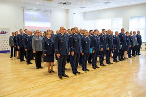 Zdjęcie przedstawia dużą grupę funkcjonariuszy Policji ustawioną w kilku równych rzędach w jasnej, nowoczesnej sali. Wszyscy są ubrani w galowe mundury policyjne. W tle widać ekran z wyświetlonymi flagami Polski i Unii Europejskiej, co sugeruje, że jest to uroczystość o charakterze oficjalnym. W sali panuje jasne, naturalne oświetlenie, a podłoga wykonana jest z jasnego drewna. Obecność kobiet i mężczyzn w różnych mundurach wskazuje na udział funkcjonariuszy z różnych jednostek lub pionów służby.