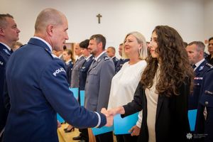 Zdjęcie przedstawia uroczysty moment wręczania gratulacji lub dyplomu przez funkcjonariusza Policji nadinsp. Romana Kustera Zastępce Komendanta Głównego Policji kobiecie cywilnej,j podczas oficjalnej ceremonii. Funkcjonariusz ubrany jest w galowy mundur policyjny z widocznym napisem „POLICJA” na ramieniu. Ściska dłoń młodej kobiecie o długich, ciemnych, kręconych włosach, ubranej w elegancki strój cywilny. Oboje uśmiechają się w geście wzajemnego szacunku.W tle widoczna jest większa grupa funkcjonariuszy, w tym kobiety i mężczyźni w różnych mundurach – zarówno granatowych, jak i szarych – co sugeruje udział przedstawicieli różnych formacji lub szczebli Policji.