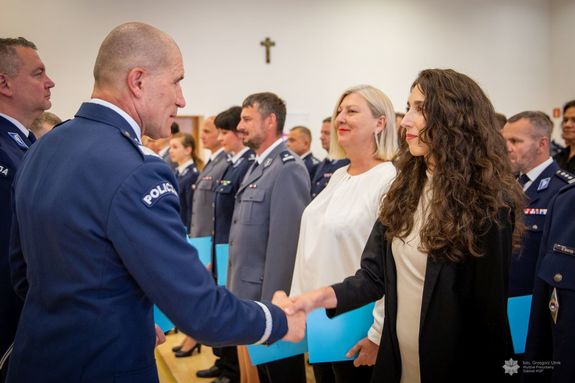 Zdjęcie przedstawia uroczysty moment wręczania gratulacji lub dyplomu przez funkcjonariusza Policji nadinsp. Romana Kustera Zastępce Komendanta Głównego Policji kobiecie cywilnej,j podczas oficjalnej ceremonii. Funkcjonariusz ubrany jest w galowy mundur policyjny z widocznym napisem „POLICJA” na ramieniu. Ściska dłoń młodej kobiecie o długich, ciemnych, kręconych włosach, ubranej w elegancki strój cywilny. Oboje uśmiechają się w geście wzajemnego szacunku.W tle widoczna jest większa grupa funkcjonariuszy, w tym kobiety i mężczyźni w różnych mundurach – zarówno granatowych, jak i szarych – co sugeruje udział przedstawicieli różnych formacji lub szczebli Policji.