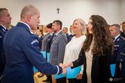 Zdjęcie przedstawia uroczysty moment wręczania gratulacji lub dyplomu przez funkcjonariusza Policji nadinsp. Romana Kustera Zastępce Komendanta Głównego Policji kobiecie cywilnej,j podczas oficjalnej ceremonii. Funkcjonariusz ubrany jest w galowy mundur policyjny z widocznym napisem „POLICJA” na ramieniu. Ściska dłoń młodej kobiecie o długich, ciemnych, kręconych włosach, ubranej w elegancki strój cywilny. Oboje uśmiechają się w geście wzajemnego szacunku.W tle widoczna jest większa grupa funkcjonariuszy, w tym kobiety i mężczyźni w różnych mundurach – zarówno granatowych, jak i szarych – co sugeruje udział przedstawicieli różnych formacji lub szczebli Policji.