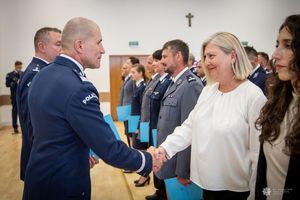 Zdjęcie przedstawia uroczysty moment wręczania gratulacji lub dyplomu przez funkcjonariusza Policji nadinsp. Romana Kustera Zastępce Komendanta Głównego Policji kobiecie cywilnej,j podczas oficjalnej ceremonii. Funkcjonariusz ubrany jest w galowy mundur policyjny z widocznym napisem „POLICJA” na ramieniu. Ściska dłoń kobiecie o jasnych włosach, ubranej w elegancki strój cywilny. Oboje uśmiechają się w geście wzajemnego szacunku.W tle widoczna jest większa grupa funkcjonariuszy, w tym kobiety i mężczyźni w różnych mundurach – zarówno granatowych, jak i szarych – co sugeruje udział przedstawicieli różnych formacji lub szczebli Policji.
