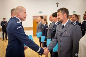 Zdjęcie przedstawia uroczysty moment wręczania gratulacji lub dyplomu przez funkcjonariusza Policji nadinsp. Romana Kustera Zastępce Komendanta Głównego Policji kobiecie cywilnej,j podczas oficjalnej ceremonii. Funkcjonariusz ubrany jest w galowy mundur policyjny z widocznym napisem „POLICJA” na ramieniu. Ściska dłoń policjantowi Centralnego Laboratorium Kryminalistycznego Policji.  Oboje uśmiechają się w geście wzajemnego szacunku.W tle widoczna jest większa grupa funkcjonariuszy, w tym kobiety i mężczyźni w różnych mundurach – zarówno granatowych, jak i szarych – co sugeruje udział przedstawicieli różnych formacji lub szczebli Policji.