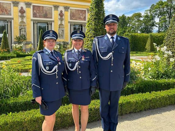 Podczas obchodów Święta Policji na tle zamku w Wilanowie stoją obok siebie  Dyrektor Centralnego Laboratorium Kryminalistycznego Policji insp. Iwoną Marcianiak - Krawczyk oraz podkom. Emilia Liwarowska, podkomisarz  Maciej Iwoła biegli Centralnego laboratorium Kryminalistycznego Policji i wspólnie pozują do zdjęcia.