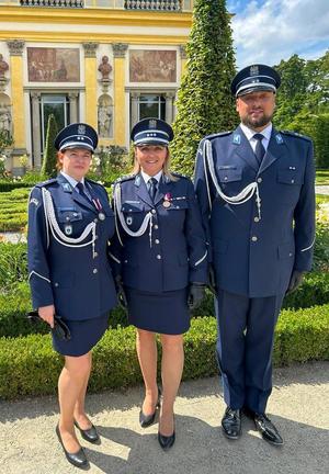 Podczas obchodów Święta Policji na tle zamku w Wilanowie stoją obok siebie  Dyrektor Centralnego Laboratorium Kryminalistycznego Policji insp. Iwoną Marcianiak - Krawczyk oraz podkom. Emilia Liwarowska, podkomisarz  Maciej Iwoła biegli Centralnego laboratorium Kryminalistycznego Policji i wspólnie pozują do zdjęcia.