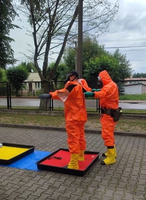 2 osoby ubrane w kombinezony zabezpieczające koloru pomarańczowego stoi obok siebie. jedna osoba stara się rozebrać i odkazić po skończonych ćwiczeniach stojąc w pojemniku z cieczą.